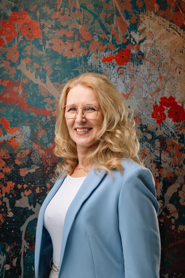 Vrouw met bril in een lichtblauwe blazer over een wit shirt, voor een geschilderd bloemendoek