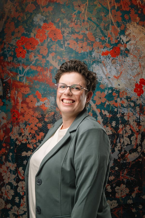 Vrouw met bril in een groene blazer, half naar de camera gedraaid, voor een geschilderd bloemendoek