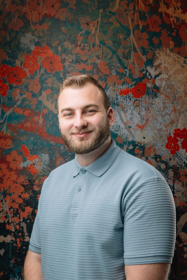 Jonge man met baard in een lichtblauwe polo, voor een geschilderd bloemendoek
