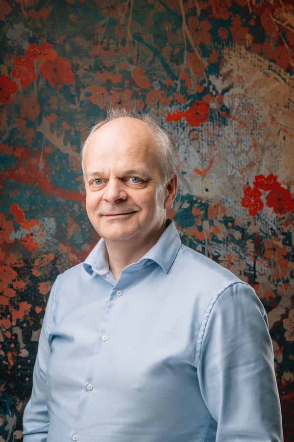 Man in een lichtblauw overhemd met opgerolde mouwen, voor een geschilderd bloemendoek