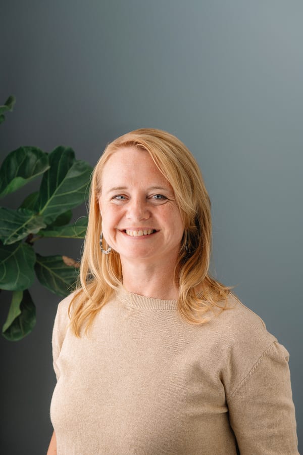 Vrouw met blond haar in een beige trui, voor een grijsgroene wand met een vijgenboom