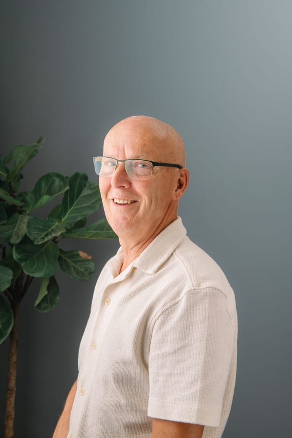 Man met bril in een crème poloshirt, half gedraaid, voor een grijsgroene wand met een vijgenboom