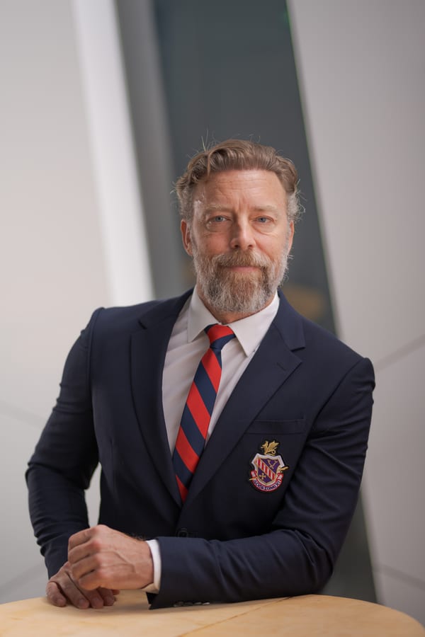 Man met grijze baard in een donkerblauw colbert met stropdas, aan een tafel, tegen een donkere achtergrond