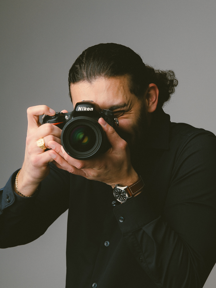Fotograaf met de camera aan het oog, tegen een effen grijze achtergrond